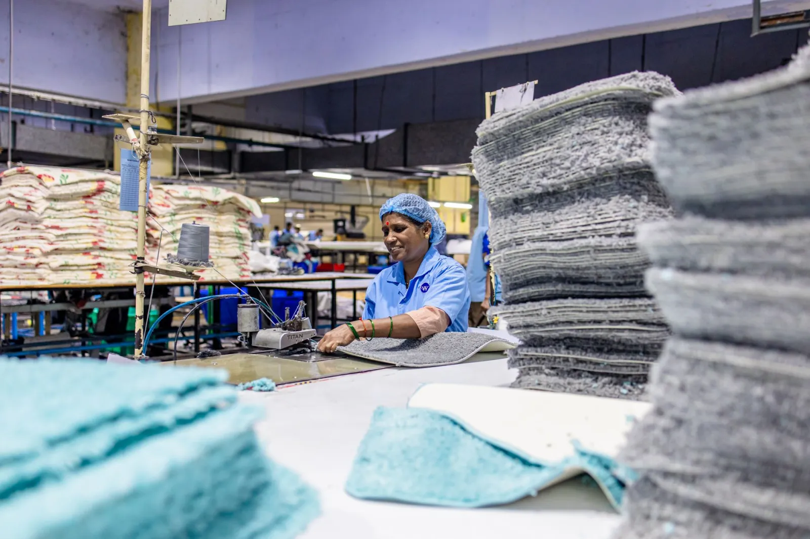 Female factory worker stitching textiles with machinery in a busy production facility.